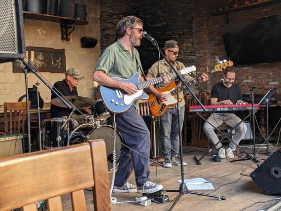 A photo of the Slim Kings playing outdoors at the LIC Bar in the afternoon. The photo is taken from the side, with a singer/guitarist, bassist, keyboardist and drummer. The drummer is Liberty DeVitto, formerly of Billy Joel's band.