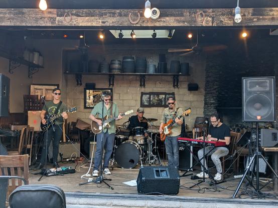 A photo of the Slim Kings playing outdoors at the LIC Bar in the afternoon. The photo is taken from the front with the full band visible: lead guitiarist, lead singer/guitarist, bassist, keyboardist and drummer. The drummer is Liberty DeVitto, formerly of Billy Joel's band.
