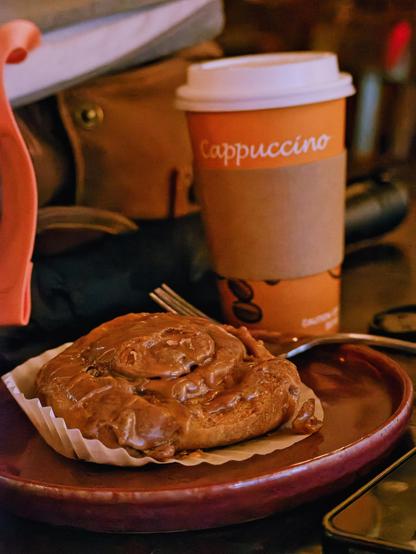 A gooey, dark caramel apple cinnamon rolls sits on a red ceramic plate sitting in front of a brown cup that reads "Cappucino" with a cardboard coffee collar. A brown bag sits just behind and the background fades to a blur.