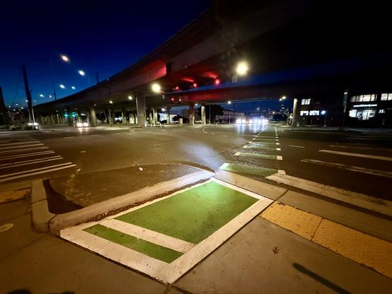 Wide angle view of the 6th Avenue bike lane crossing at Spokane Street underneath a viaduct; the bike lane continues across the intersection passing under the viaduct