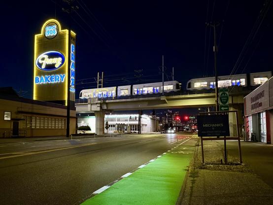 A green painted flexpost protected bike lane extends on 6th Avenue near a tower lit with neon lights for Franz Bakery as a light rail train passes by on elevated tracks at night