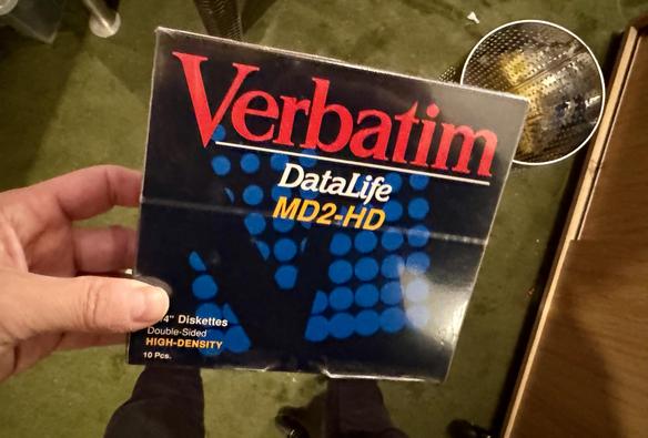 A hand is holding a box of Verbatim DataLife MD2-HD 3.5-inch diskettes, labeled as double-sided and high-density, containing 10 pieces. The background features a patterned floor and a metallic waste basket.
orginalverpacket