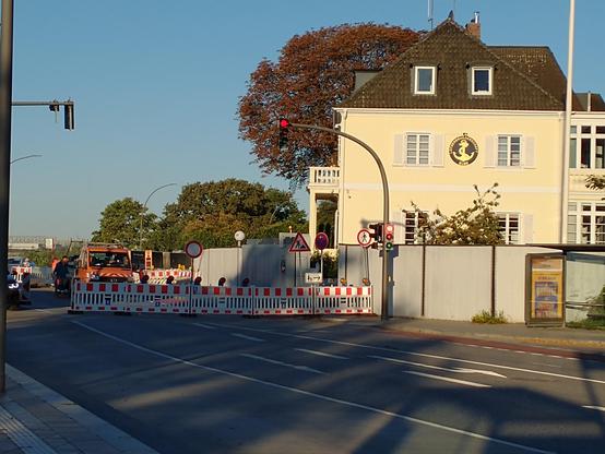Das Bild zeigt eine Straße, an der Fußgängerampel steht eine rot-weiße Baustellenabsicherung quer über die Fahrbahn, die Durchfahrt ist verboten. Im Hintergrund steht ein hellgelb gestrichenes Haus und einige Bäume.