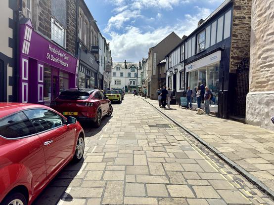 Sunlight spills along High Street in Conwy, where stone-paved streets wind between historic buildings and small independent shops. A bright purple storefront for St David’s Hospice adds a pop of color beside traditional stone facades, while a mix of modern and classic cars line one side of the narrow lane. Opposite, people pause outside a shop with wide windows, some chatting, others accompanied by a resting dog, as the street gently rises toward whitewashed buildings framed by a blue sky with scattered clouds. The scene blends medieval charm with everyday life, capturing the quiet rhythm of a Welsh market town.