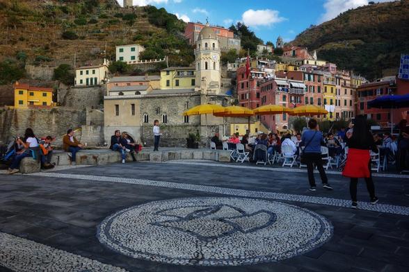 Lively scene of Vernazza’s central square in Cinque Terre, Italy, showcasing the village’s iconic charm. The square is adorned with a large, decorative pebble mosaic at its centre. People are gathered at outdoor seating under vibrant yellow umbrellas, enjoying food and conversation. The backdrop features the village’s signature pastel-coloured buildings—yellows, pinks, and oranges—stacked along the terraced hillside, with historic stone structures and a prominent church tower. The atmosphere is bustling, with locals and tourists mingling, while the surrounding green hills add to the scenic beauty of this coastal gem.