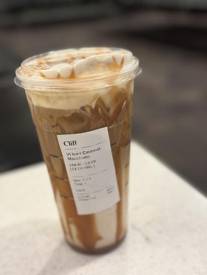 The image shows a clear plastic cup filled with an iced beverage, likely a caramel macchiato, with a creamy texture and visible ice cubes. The drink is topped with a layer of foam and caramel drizzle. A white label on the cup reads "Cliff" at the top, followed by "Vt Iced Caramel Macchiato" and "VAN SC COLD FM [EX CARMAL]" in smaller text. The label also indicates "Item 1 of 1" and "Total 1," suggesting it is a single item order. The cup is placed on a light-colored surface, possibly a table, with a blurred background that suggests an indoor setting.