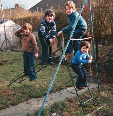 A scan of a colour, square photograph taken around 1980. We're at the bottom end of a garden with short grass on it separated by a narrow concrete path. At this bottom end we can see a section fenced off in which bushes and compost is left and on the left of the view we can see the backs of houses, a shed, and separated by a wire fence from us there's the tail end of a neighbouring garden too. The main focus is on the four young boys and the equipment down here well away from the house. A metal swing frame is to the right, mostly out of view, just a couple of A-frame supports visible with chains for a swing but lacking any seat. Arranged perpendicular to it is a child's slide. Again, it's just a metal frame in an angled L-shape, with no sliding plastic area and just thin metal, rusted rungs in each part of it. The four young boys are arranged all over it. Two are on the slide section of the slide equipment, one is at its apex but resting back towards the adjacent swing frame, and one is climbing the slide steps but gripping the chains of the swing nearby. All four are looking at the camera and grinning. All four wear dark jeans and thick jumpers of varying styles. The fashion, the browning of the grass, the lack of leaves on the bushes, the overall muted colours suggest it's late or early in the year and a little chilly.