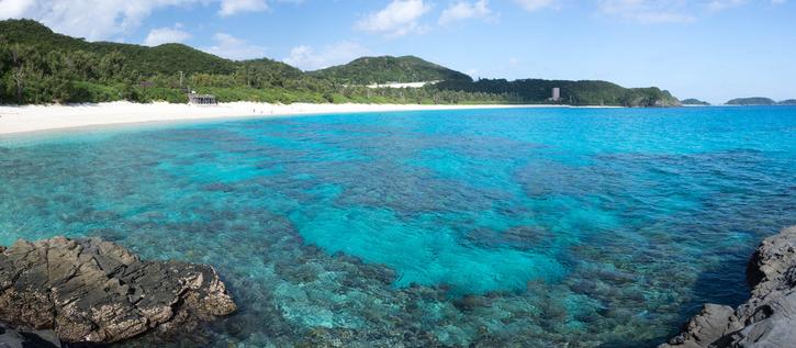 A panoramic view of Zamami Island in Okinawa, Japan, showing turquoise-blue waters with coral visible beneath the surface, a stretch of white sandy beach lined with lush green forest, and rocky outcrops in the foreground under a bright blue sky.