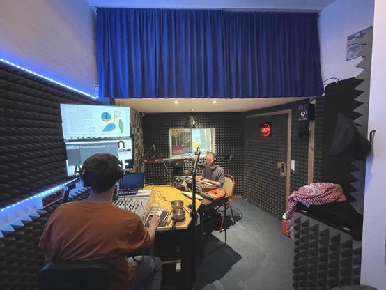 The studio is occupied by a figure at the mixer reading from a monitor and a figure playing the keyboard set slightly further back into the room. A blue curtain adorns the upper field of view.