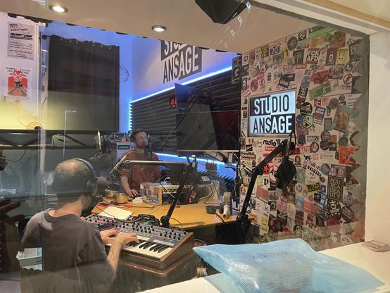 The studio is now seen from the reverse angle, through the glass that separates the recording room from the public. We see the reverse of each person, reading and playing.