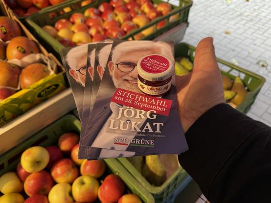 Flyer und ein Marmeladenglas mit Aufdruck „Stichwahl am 28. September – Jörg Lukat“ vor Apfelkisten auf einem Marktstand.