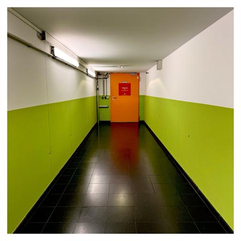 Quadratisches Foto. Blick durch den Gang eines Parkhauses. Der Boden ist schwarz gefliest, die Wände sind in der unteren Hälfte hellgrün, die obere Hälfte und die Decke sind weiß. Am Ende des Ganges ist eine orangefarbene Türe, darauf ist ein rotes Schild. Darauf steht Tiefgarage erstes Untergeschoss. Die führenden Linien im Bild starten genau in den Ecken, wodurch alles symmetrisch ist.
