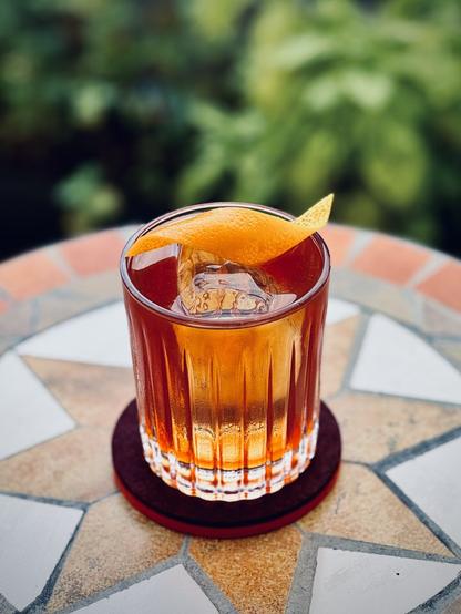 a reddish-brown cocktail with orange twist garnish and big rock of clear ice sitting on a tile table with plants in the background