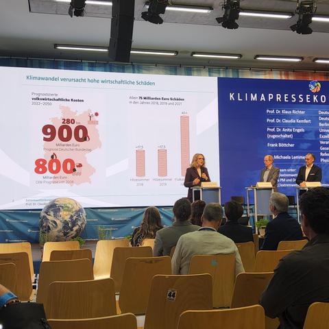 Claudia Kemfert auf der Pressekonferez zum Klimaaufruf der DPG und DMG auf dem Extremwetterkongress. Im Hintergrund eine Folie, die Volkswirtschaftliche Kosten der Klimakrise für Deutschland zeigt.