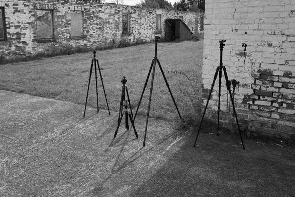 B&W-Aufnahme von fünf Kamerastativen. Diese stehen draußen zwischen zwei Baracken-Ruinen der Gedenkstätte.