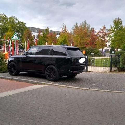 Schwarzes Luxus-SUV steht auf der Zugangsfläche vor einem Kinderspielplatz in einem verkehrsberuhigten Bereich.