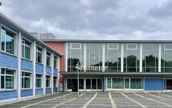 Farbiges Querformat. Bahnhofsgebäude im sachlichen Baustil unter bewölktem himmel.
