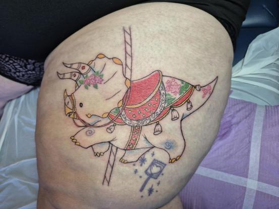 Photo of an in progress tattoo, with some of the colours done. It is of a happy chonky triceratops as a carousel ride!