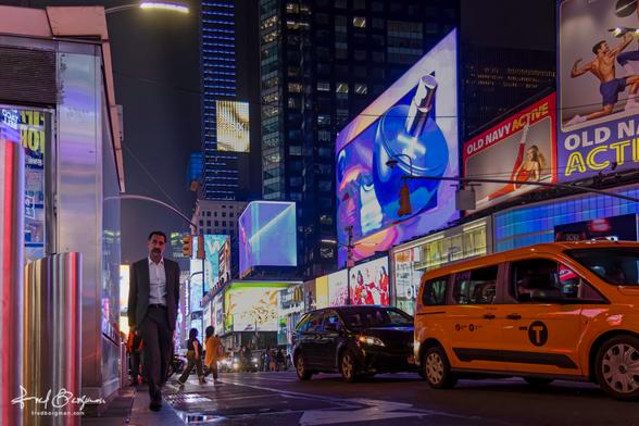 Man in suit walking away from Times Square at night with passing cars in the street. The buildings are full of digital advertising signs.