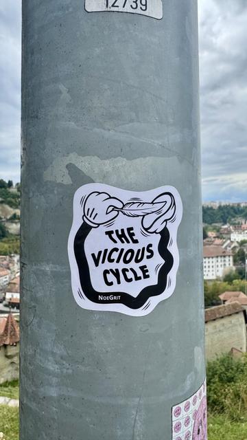Sticker on a pole representing a cartoonish hand linked to foot by a black limb and is ticking the foot with a feather. The text on the sticker says «  The Vicious Cycle » by NoeGrit