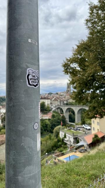 Sticker on a pole representing a cartoonish hand linked to foot by a black limb and is ticking the foot with a feather. The text on the sticker says «  The Vicious Cycle » by NoeGrit