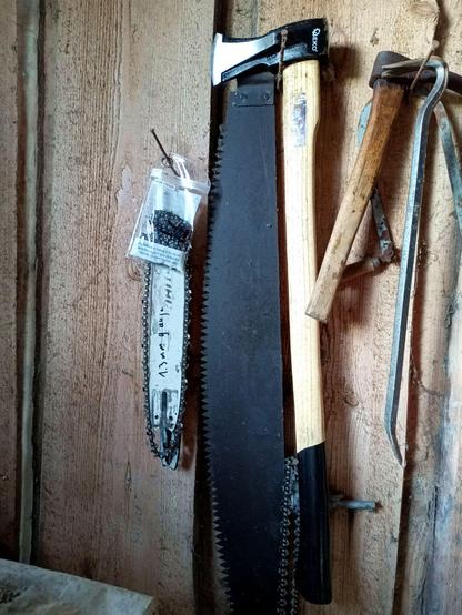 A bunch of stuff hanging on a garbarn wall. Center is a newish looking splitting axe with Geko logo on the head and a wooden handle with black painted end. The label is peeling off, otherwise it looks shiny and nice with mild signs of use despite having chopped through a considerable amount of firewood. There's a chainsaw chain and bar hanging there, an old two person felling saw, an adze and a crowbar.