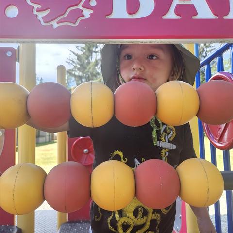 A young child wearing a hat looks through a playground abacus, its colorful spheres alternating red, yellow, and positioned below a pink and white sign.