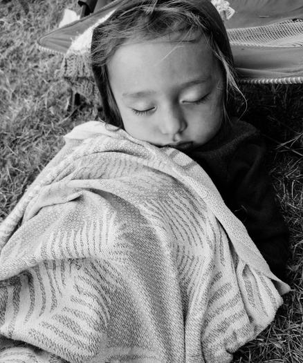 Black and white image of a young child sleeping outdoors on grass, wrapped in a patterned blanket, with eyes closed and head resting on the blanket.