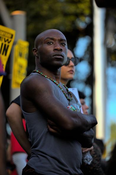 A Black man standing at a protest, looking above and beyond the camera. His arms are crossed and is looking intently towards a speaker. Behind him is more of the crowd holding anti-war signs.