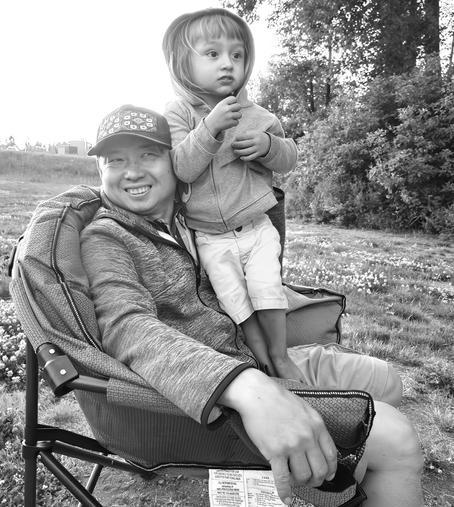 Black and white image of a man sitting in a camping chair with a young child standing on his lap; the man wears a baseball cap, a zip-up hoodie, and shorts, smiling at the camera while the child wears a hooded jacket and shorts, looking to the right while holding an item; they are in a grassy field with trees in the background.