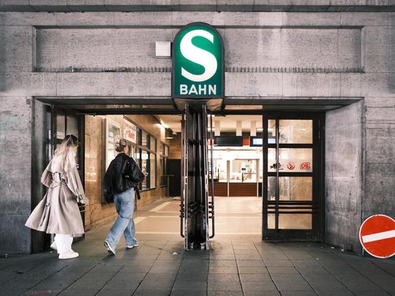 Das Bild zeigt den Eingang eines S-Bahn-Bahnhofs. Zwei junge Frauen gehen gerade in den Bahnhof hinein. Der Eingang ist aus Beton und wirkt etwas in die Jahre gekommen. Drei von vier Türflügeln sind geöffnet. Über dem Eingang befindet sich das grüne S-Bahn-Schild. Rechts neben dem Eingang steht ein rotes "Einfahrt verboten"-Schild an die Wand gelehnt. Im Hintergrund ist der Innenraum des Bahnhofs zu sehen. The picture shows the entrance to an S-Bahn station. Two young women are just entering the station. The entrance is made of concrete and looks a little worn. Three of the four wings of the doors are open. Above the entrance is the green S-Bahn sign. To the right of the entrance, a red “No Entry” sign is leaning against the wall. The interior of the station can be seen in the background.