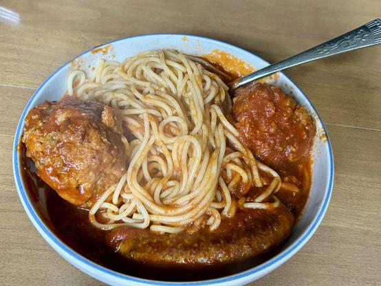 A large bowl of spaghetti with meatballs for eyes and a hot sausage for a smile with a fork in the upper right. The bowl sits on a brown, shining table.