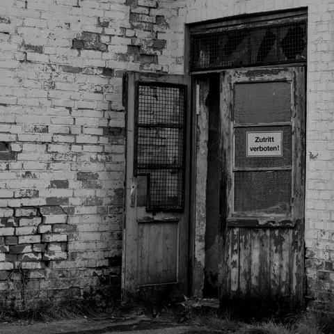 Eine alte Holztür an einer Stein Baracke in der Lagergedenkstätte.
Ein Flügel der Tür ist ganz auf und der andere fast geschlossen. Alles sehr marode und kurz vorm zusammenfallen. An der Tür hängt ein Schild mit der Aufschrift betreten verboten