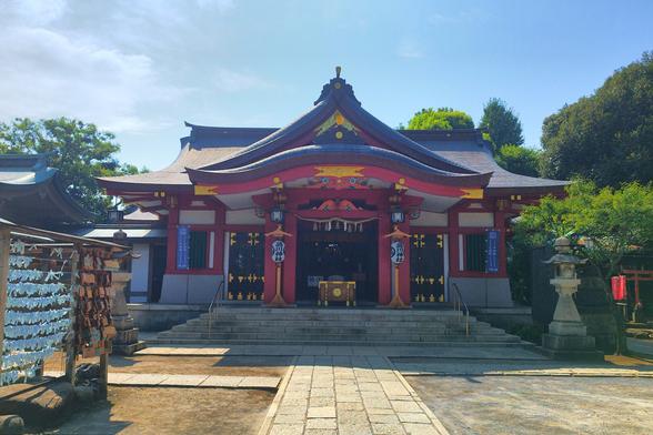 Lovely big shrine atop a hill. Built from wood, the timeframes painted bright red, everything else white. There are brass highlights as well. The roof is dark turquoise. The sun is behind it. In the shade in front of the shrine are two stone lanterns. The left one is partially obscured by the rack for wishes/horoscopes being tied after reading. A handful of trees in the background and almost perfect blue sky.