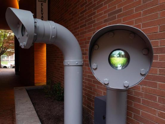 In a brick-lined planter along a quiet breezeway, two periscope-like grey pipes erupt from the ground like brontosauruses. They each are painted industrial grey and are roughly person-sized. From this angle, the closer "Periscope" reveals a bulbous screen that is displaying a watery natural scene.