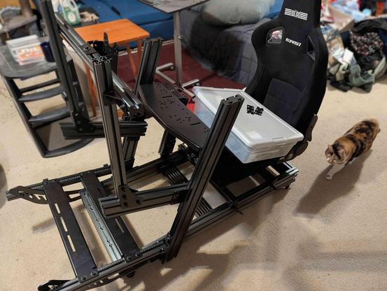 A black aluminum-extrusion racing simulator rig is partially assembled on a beige carpet. The rig includes the base frame, uprights for the wheel deck and pedal plate, and a high-backed, black racing seat (ERSI model) mounted on the back. A clear plastic storage bin rests on the wheel deck section. A calico cat walks away from the rig toward the right side of the image. The background shows a slightly messy living space with a blue couch, a small wooden table, and other items.