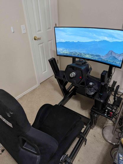 A fully assembled black aluminum-extrusion racing simulator rig is set up in the corner of a carpeted room. The rig features a deep black racing bucket seat (seen from the back), a wheel deck holding a Logitech steering wheel base, and a curved monitor mounted on a separate stand displaying a scenic view of mountains and sky. To the right of the wheel, a side mount holds a shifter and possibly a handbrake. The rig is positioned near a white closed door.