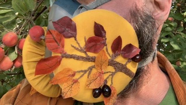 Me sporting a elastomeric mask slipcover diptych, left side. Yellow background with a branch of Cotoneaster hedge: multi-toned embroidery branches and silk leaves attached at the stem only, dark red leaves at top, transitioning to orange, and yellow at the bottom. Purple glass pendant beads act as the autumn berries on common Edmonton variants of this plant. 

(Right side in other photo: was unable to add alt text to that image)