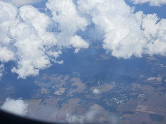 View from a plane of the ground, a mix of brown and green geometric shapes and some curves, above a couple of white clouds.