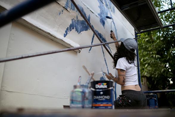 A young female artist sits on scaffolding, painting a blue mural on a white background. She holds a paint roller in her right hand and a phone in the other. The frame of the photo places us inside the scaffolding with its metal bars and paint pots scattered here and there.