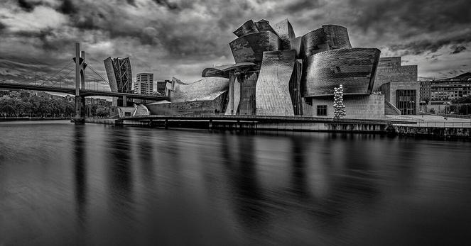 Modern architectural building with a striking design, featuring metallic, curved shapes and reflective surfaces. Positioned alongside a calm river with a smooth water effect. A bridge extends across the left side, while cloudy skies form the backdrop. An arrangement of circular art pieces stands near the structure.