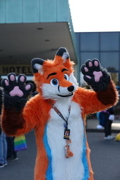 An orange fox fursuiter (ToasterTheFox) in front of an hotel entrance with his paws in the air