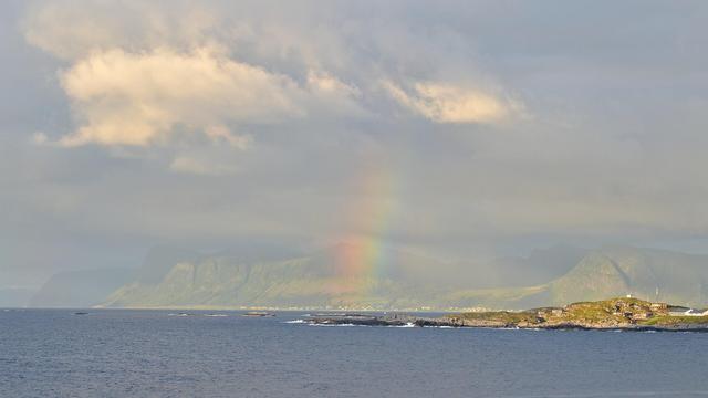 A photo of a rainbow in front of green mountains going into the sea. There's a small peninsula in front of the mountain shoreline with some buildings on it.  The sky is cloudy with some clouds being lit up by the sun.