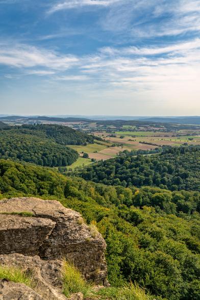 Das Bild zeigt eine weite Landschaftsaufnahme, aufgenommen von einem erhöhten Standpunkt, wahrscheinlich einem Felsvorsprung oder einer Klippe, die im Vordergrund markant zu sehen ist.
Vordergrund und Perspektive
Der Vordergrund wird von einer massiven, zerklüfteten Felsformation dominiert, die links unten ins Bild ragt. Die Textur des Steins ist rau und detailliert, mit etwas Gras, das an seinen Rändern wächst. Diese Position bietet eine tolle Aussicht auf die dahinterliegende Szenerie.
Mittel- und Hintergrund
Direkt unterhalb des Aussichtspunktes fällt der Blick in einen dichten, üppig grünen Laubwald, der sich über Hügel und Täler erstreckt. Die Baumkronen zeigen eine reiche Palette an Grüntönen und bilden einen starken Kontrast zum Stein im Vordergrund.
Der Mittelgrund öffnet sich zu einer sanft gewellten Kulturlandschaft. Hier wechseln sich Wälder mit Feldern und Wiesen ab, die in verschiedenen helleren Gelb-, Braun- und Grüntönen erscheinen, was auf landwirtschaftliche Nutzung hindeutet. Man kann vereinzelte Baumgruppen und Hecken erkennen, die die Felder gliedern.
Im Hintergrund verschwimmen die Konturen der Landschaft in einem leichten Dunst. Es sind weitere bewaldete Hügel und Höhenzüge zu sehen, die sich bis zum Horizont erstrecken und die Weite der Region betonen.
Licht und Atmosphäre
Das Bild wurde bei klarem Wetter und hellem Tageslicht aufgenommen. Der Himmel ist weit und blau, durchzogen von zarten, weißen Schleierwolken, die für eine dynamische Textur sorgen. 