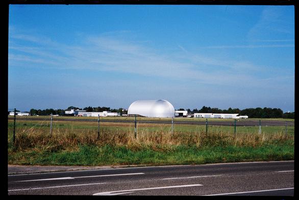 A silver airship hangar with a rounded dome-like form rises against a blue sky, surrounded by low industrial buildings on a flat airfield. A wire fence and grassy verge line the roadside in the foreground, while the hangar gleams in the midday sun. Black 35 mm film border visible. (Photo by me, CC-BY-4.0, Adox Color Mission @ EI 160, Zeiss Sonnar 50 mm f/2 1950s)