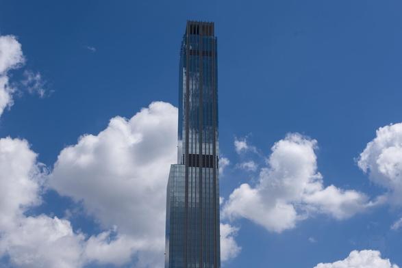 The image captures the grandeur of a towering skyscraper under a vast, clear sky dotted with fluffy clouds. The building's glass facade reflects its surroundings, giving it an almost ethereal quality as if it were suspended in air. Its rectangular form is punctuated at intervals by black horizontal stripes that likely represent floors or structural divisions.

The perspective from which the photo was taken allows for an unobstructed view of both the building and the expansive sky behind it. The cloud formations in various shades of white suggest different altitudes or types of clouds; some are large and fluffy, indicating fair weather conditions at that time, while others appear smaller and more dispersed.
