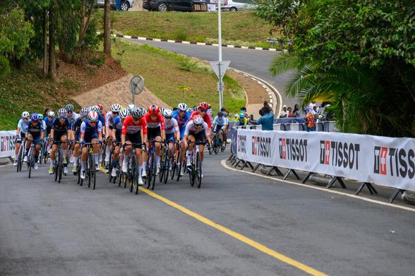A group of female cyclists in competitive gear is racing on a paved road. They are surrounded by greenery and a banner in the background. Each cyclist is focused on the race, showcasing various team colors and helmets.