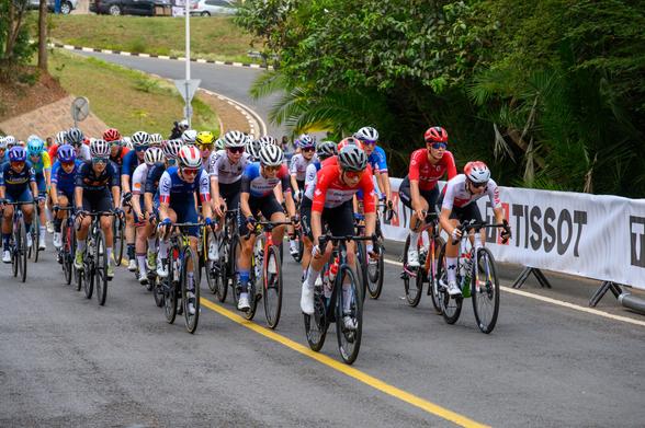 A group of female cyclists in competitive gear is racing on a paved road. They are surrounded by greenery and a banner in the background. Each cyclist is focused on the race, showcasing various team colors and helmets.