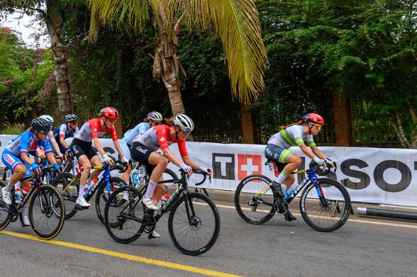 A group of female cyclists in competitive gear is racing on a paved road. They are surrounded by greenery and a banner in the background. Each cyclist is focused on the race, showcasing various team colors and helmets.