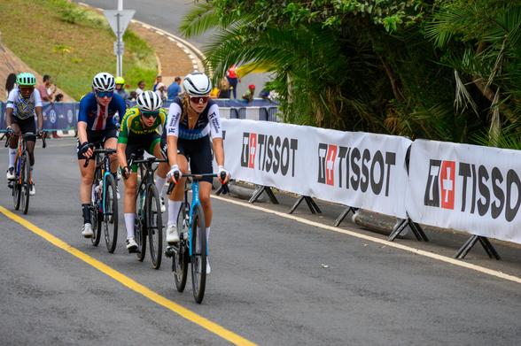 A group of cyclists in competitive attire are riding in a race, seen from behind. They are navigating a curved road, with multiple bicycles in motion showing various colors and styles. The setting appears outdoors with trees in the background.